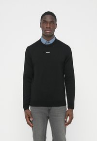 Pulover negru tricotat cu gât rotund, cu logo-ul alb „HUGO” pe piept. Asortat cu o cămașă cu guler azul deschis pe dedesubt.