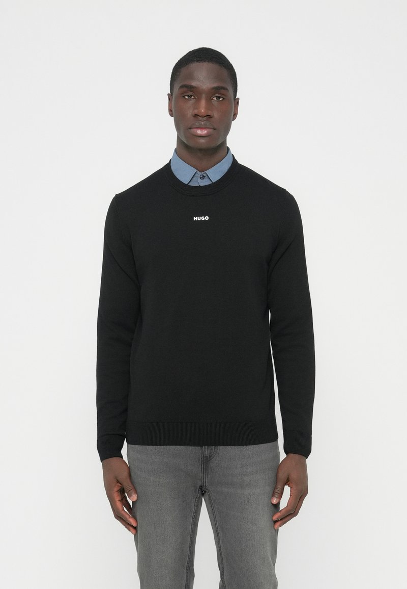 Pulover negru tricotat cu gât rotund, cu logo-ul alb „HUGO” pe piept. Asortat cu o cămașă cu guler azul deschis pe dedesubt.