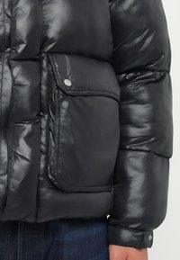 Chaqueta de plumas negra con un diseño brillante y acolchado, con un bolsillo frontal y cierre de botones. El puño tiene un detalle de botón.