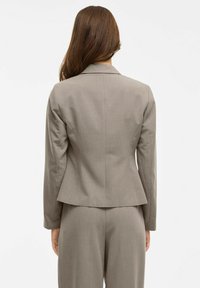 Šedý přizpůsobený blazer s dlouhými rukávy, strukturovanými rameny a seštípnutým pasem, který se vyznačuje čistým, minimalistickým designem a hladkým povrchem.
