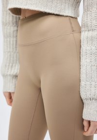 Κοντινό πλάνο ατόμου που φοράει μπεζ κολάν με ψηλή μέση και κρεμ πλεκτό πουλόβερ, παρουσιάζοντας λεπτομερείς ραφές και υφή.