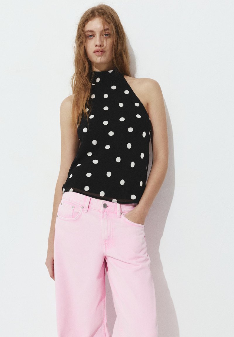 Mlada žena s dugom svijetlosmeđom kosom, odjevena u crni top na vezanje s bijelim točkicama i visokim svijetloružičastim hlačama, stoji uz bijeli zid.