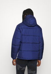 Mörkblå puffjacka med huva, med horisontellt kviltat mönster och elastiska muddar. Slät, glänsande textur, lättviktig och isolerad.