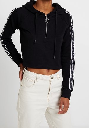 Sort cropped hoodie med lynlås foran, snor og metalring til at trække i. Har hvide side-striber med gentaget tekst. Parret med lyse bukser.