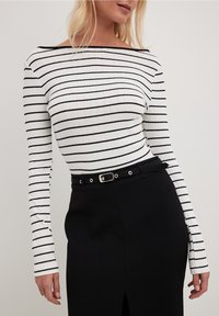 Vit långärmad topp med tunna svarta horisontella ränder, ribbad textur och bred båtformad halsringning, kombinerad med en svart kjol med ett silverfärgat bälte.