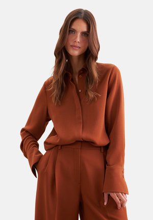 Femme aux longs cheveux bruns portant une chemise boutonnée couleur rouille et un pantalon taille haute assorti, une main dans la poche.