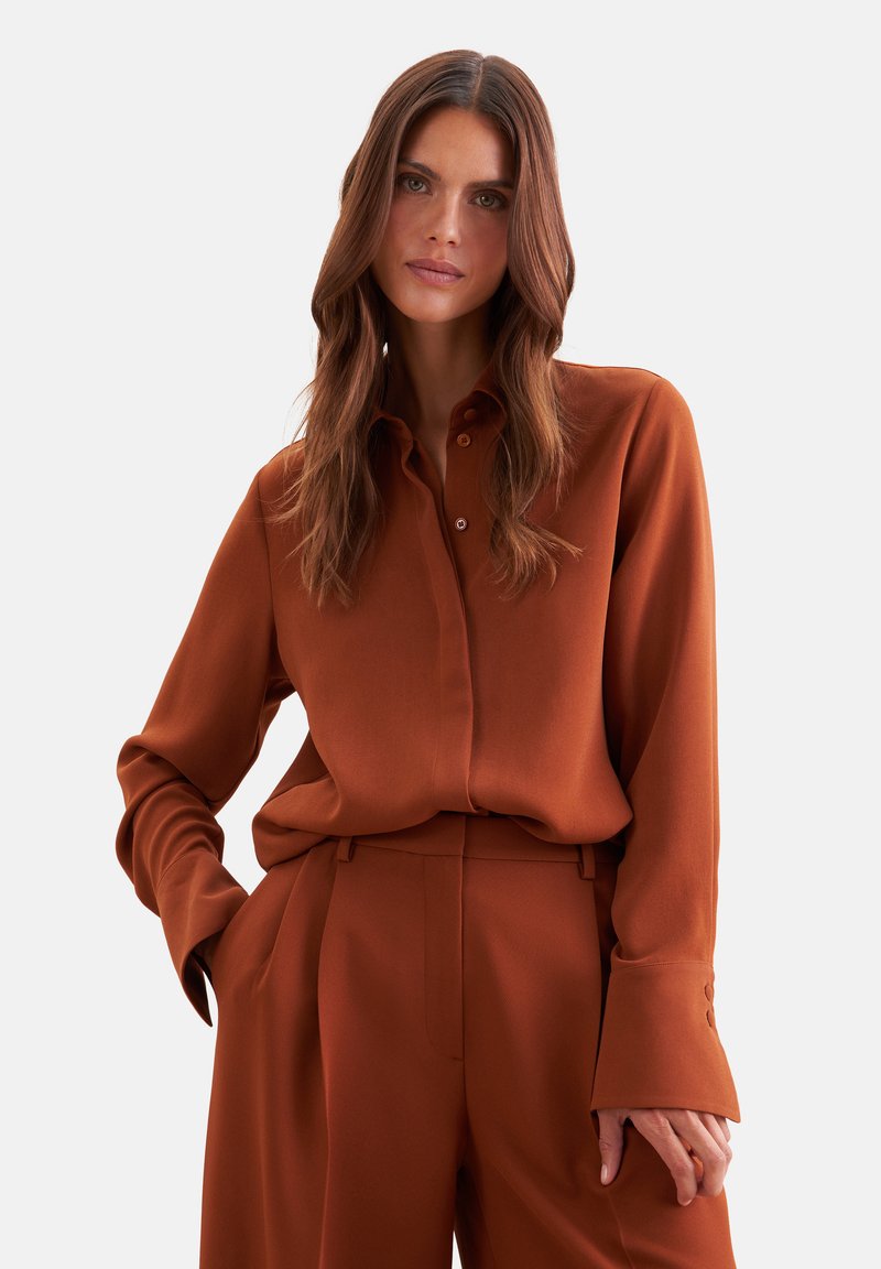 Femme aux longs cheveux bruns portant une chemise boutonnée couleur rouille et un pantalon taille haute assorti, une main dans la poche.
