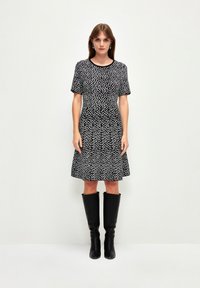 Schwarzes Kleid mit Rundhalsausschnitt, kurzen Ärmeln und silhuette im Fit-and-Flare-Stil. Hat ein weißes abstraktes Muster. Trägt dazu knielange schwarze Stiefel.