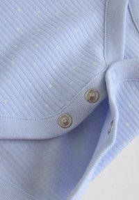 Indumento per bambini azzurro chiaro con texture a costine sottili, piccoli puntini bianchi e due bottoni a pressione argentati parzialmente chiusi.