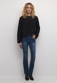 Melnā, puscaurspīdīgā kapucē ar perforētu dizainu, priekšējo kabatu un auklu. Apvienota ar gaiši zilām taisnās kājas džinsām un melnām potīšu zābakiem.