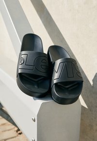 Sandales à enfiler noires en matériau synthétique avec une bride ajustable texturée ornée d'un logo Levi's en relief. Design minimaliste.