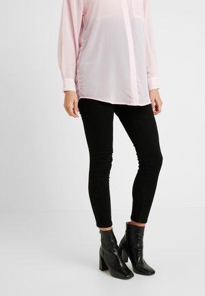 Osoba ubrana w jasnoróżową koszulę na guziki, czarne dopasowane spodnie oraz czarne botki na obcasie, stojąca na jednolitym tle.