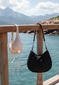 Μπεζ πάνω μέρος μπικίνι και μαύρη τσάντα κρεμασμένα σε ξύλινη περίφραξη δίπλα στον ωκεανό, με βραχώδη ακτογραμμή και βουνά στο φόντο κάτω από γαλάζιο ουρανό.