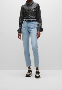 Chemise en cuir synthétique noir avec deux poches poitrine, jean bleu clair à jambes droites, et baskets noires avec des accents beige et blanc.