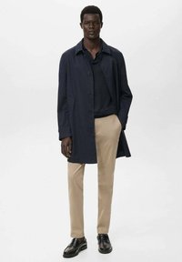 Lange marineblauwe jas met knoopsluiting, gecombineerd met een donkere top en beige broek. Zwarte schoenen maken de outfit compleet. Gladde stoftextuur.