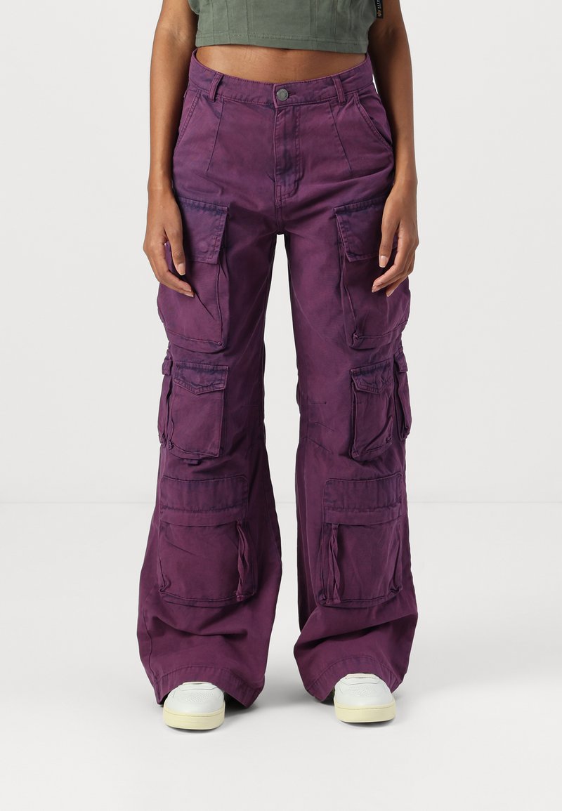 Paarse cargobroek met een loose fit, meerdere zakken en versterkte naden. Materiaal lijkt textuur te hebben. Ggedragen met witte sneakers.