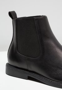 Bottine en cuir noir avec des panneaux élastiques sur les côtés, une texture lisse, un bout rond et une semelle en caoutchouc plate et basse.