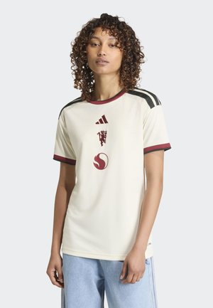 Kvinde med krøllet hår bærer en cremefarvet Adidas-shirt med sorte striber, bordeaux krave og logoer, kombineret med lyseblå jeans.