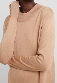 Pullover beige con collo alto, maniche lunghe e polsini a coste. Presenta bottoni decorativi dorati su una manica, realizzato in materiale morbido.