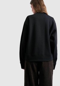 Femme aux longs cheveux portant un sweat-shirt noir surdimensionné et un pantalon sombre, vue de dos sur un fond uni gris clair.