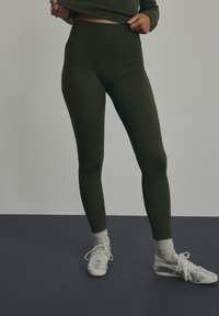 Olivgrüne, gerippte Leggings mit hohem Taillendesign, kombiniert mit silbernen Sneakern und weißen Sneakersocken. Schlichte Details und glatte Textur.