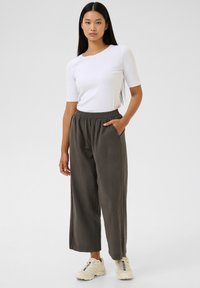 Balti trumpomis rankovėmis megztinis derinamas su laisvomis tamsiai žaliomis, su elastiniu juosmeniu kelnėmis. Avalynė: smėlio spalvos sportbačiai; modelis stovi neutraliame fone.