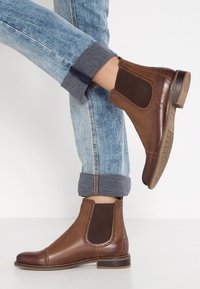 Brązowe skórzane botki za kostkę z elastycznymi panelami po bokach, kontrastowym przeszyciem oraz niskim obcasem, zestawione z podwiniętymi jasnoniebieskimi dżinsami.