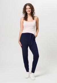 Top con spalline rosa chiaro, pantaloni navy con cintura annodata e sneakers bianche. Modella in piedi con capelli ricci, mani in tasca, sfondo neutro.