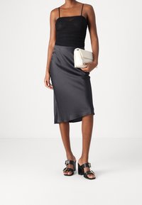 Schwarzes, gerafftes Tanktop mit dünnen Trägern, kombiniert mit einem dunkelgrauen, satinierten Rock. Schwarze Sandalen mit Blockabsätzen und eine weiße Clutch in der Hand.