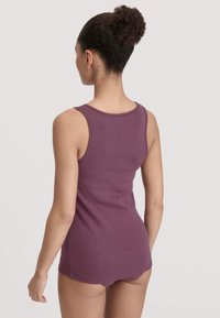 Vrouw met krullend haar in een bun, gekleed in een mouwloze paarse top en bijpassend ondergoed, met haar rug naar de camera gekeerd tegen een effen achtergrond.