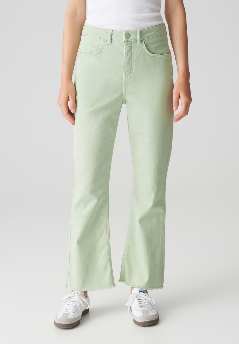 Jeans a zampa verde chiaro con tasche frontali, chiusura a bottone e orlo sfilacciato. Indossati con scarpe da ginnastica bianche, che mostrano un look casual.