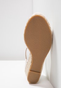 Semelle d'une espadrille compensée beige avec un grip en caoutchouc texturé, présentée en position verticale contre un coin de mur blanc.