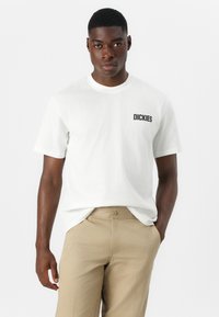 Camiseta blanca de algodón con un logo negro de "DICKIES" en el lado izquierdo. El modelo la combina con unos pantalones beige. Diseño simple de cuello redondo.