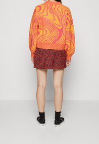 Kvinna iklädd en rosa och orange mönstrad tröja, röd och svart mini kjol med hjärttryck, svarta strumpor och svarta slipskor stående med ryggen mot.