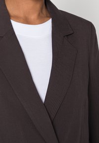 Hnědý blazer s hladkým povrchem a klopami, přehozený přes bílé tričko s kulatým výstřihem. Detailní záběr na límec a horní část.
