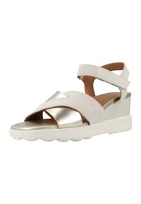 Sandalias de cuña de ante beige con una tira cruzada metálica dorada, correa de tobillo ajustable y una suela blanca gruesa con un patrón en forma de ola.