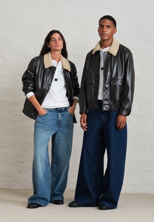 Une femme et un homme portent des vestes en cuir à col noir en peau lainée, des hauts blancs et des jeans bleus amples devant un mur de studio uni.