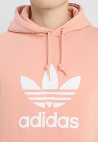 Rosa sweatshirt med dragsko och huva, med en vit Adidas-logotyp och tre lövdesign. Tillverkad av mjukt, lätt tyg.