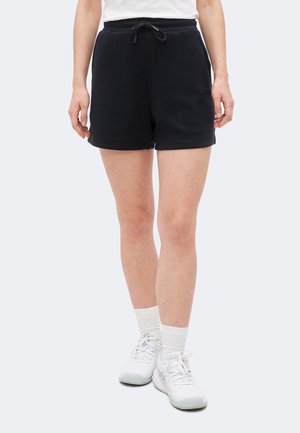 Noge koje nose crne šorc s remenom, bijele čarape i bijele sportske cipele, na jednostavnoj svijetloj pozadini.