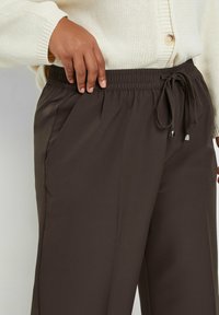 Bruine, elastische broek met een trekkoord en zilveren accenten, voorzien van zijzakken en een gladde textuur.