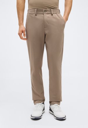 Mann trägt beige Hose und weiße Golfschuhe, steht mit einer Hand in der Tasche vor einfarbigem Hintergrund.