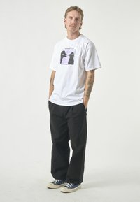 White cotton t-shirt with a graphic print of two birds against a lavender background. Paired with black trousers and blue sneakers.