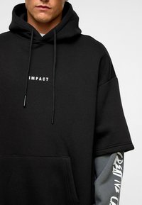 Zwarte oversized hoodie met een trekkoord en een front pocket, met de tekst "IMPACT" in het wit. Lange grijze mouwen met witte graphics.