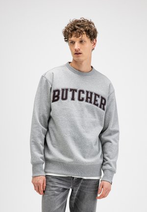 Jonge man met krullend haar die een grijze trui draagt met "BUTCHER" in grote letters, staand tegen een effen witte achtergrond.