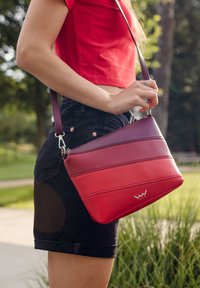 Červená a burgundská pruhovaná taška přes rameno s hladkou texturou, vybavená stříbrnými doplňky a logem. Nositelná s černými šortkami v venkovním prostředí.