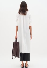 Langes weißes Hemd mit hochgekrempelten Ärmeln, kombiniert mit schwarzen Leder-Culottes. Dunkelbraune Tragetasche in einer Hand gehalten. Glatte Textur, minimalistisches Design.