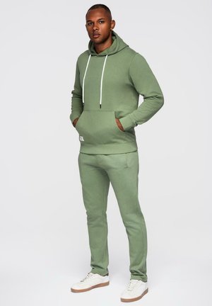 Man staat met handen in de zakken, draagt een lichtgroene capuchontrui en bijpassende broek, en witte sneakers tegen een effen achtergrond.