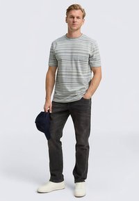 Majica s kratkimi rokavi z belimi in modrimi črtami, kombinirana s temnimi jeans hlačami in belimi supergami. Moški drži mornarsko modro kapo.