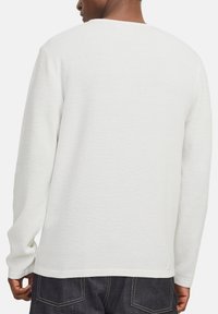 Mann, der ein strukturiertes weißes Langarmshirt und dunkle Jeans trägt, von hinten vor einem schlichten Hintergrund gezeigt.