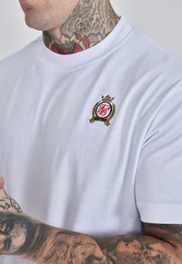 Bijela pamučna majica s okruglim izrezom, s malim vezenim logotipom u crvenoj i zlatnoj boji, prikazana na teksturiranoj površini.